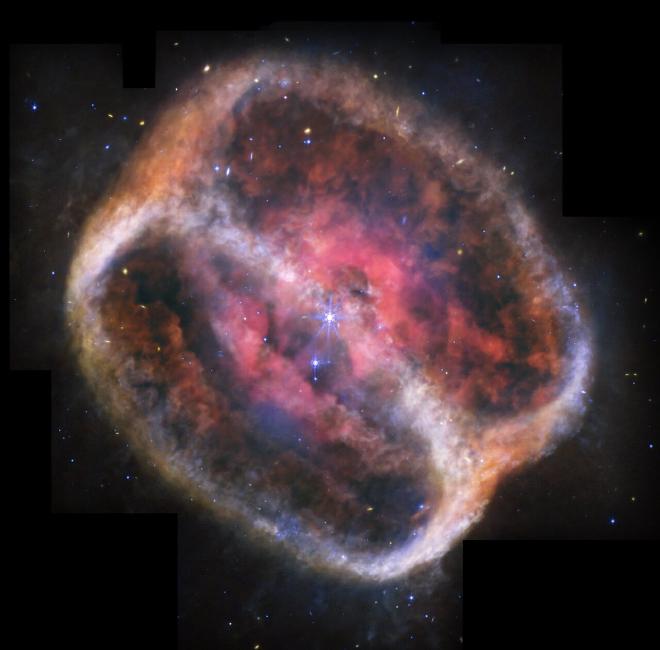What looks like a single large, bright star (but is two) shines with bright purple diffraction spikes at the center of a large, diffuse cylinder of gas and dust that is tipped to the left. At the center is a bright pink clumpy cloud that takes up about 25% of the view. There are two large rings seen at a roughly 60-degree angle that appear joined at top left and bottom right. The edges are denser, and form shallow V-shapes that go inward. The rings appear orange at top left and bottom right, and are blue at bottom and center right. There is diffuse orange material around the body. The black background of space is speckled with tiny stars and galaxies mostly in blues and yellows. Areas Webb did not observe are along the top edges, a thin vertical near the nebula at top left, and at the bottom left and right corners.Credit: NASA, ESA, CSA, STScI, Michael Ressler (NASA-JPL), Dave Jones (IAC)