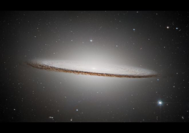 La Galaxia del Sombrero es un disco oblongo de color blanco pálido con un núcleo brillante. Parece casi de canto, pero está ligeramente inclinada al frente, lo que ofrece una vista ligeramente cenital de la región interior de la galaxia y su brillante núcleo. El disco exterior es más oscuro, con tonos marrones y negros. Galaxias distantes de diferentes colores y diversas estrellas se motean sobre el fondo negro del espacio que rodea la galaxia. Crédito de la imagen: ESA/Hubble y NASA, K. Noll LICENCIA CC BY 4.0 INT o Licencia estándar de la ESA (El contenido puede utilizarse bajo cualquiera de las dos licencias)