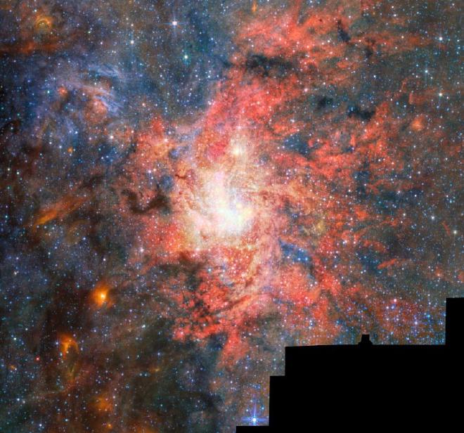 Un cúmulo estelar brillante y densamente poblado se encuentra cerca del centro de la imagen, rodeado de intrincadas nubes de gas y polvo incandescentes en tonos rojos, rosas, naranjas y azules. Franjas oscuras de polvo serpentean a través de la nebulosa, contrastando con la luminosa región central. Innumerables estrellas se encuentran dispersas por el campo, con algunas estrellas más brillantes en primer plano que muestran los característicos picos de difracción del telescopio Webb. Una región negra irregular ocupa la esquina inferior derecha de la imagen, indicando un área donde no se disponía de datos de observación. Crédito de la imagen: ESA/Webb, NASA y CSA, F. Peißker, F. Yusef-Zadeh, NB Sabha, C. Chan