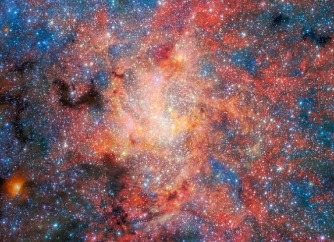 Un denso campo estelar llena toda la imagen, con innumerables estrellas dispersas sobre un fondo de nubes brillantes en tonos rojos, naranjas, rosas, blancos y azules. En el centro, una brillante concentración de estrellas crea un núcleo luminoso, casi blanco, rodeado de intrincados filamentos y volutas de gas y polvo iluminados que irradian hacia afuera. Manchas oscuras e irregulares de polvo opaco interrumpen las nubes brillantes, particularmente hacia el lado izquierdo de la imagen, donde aparecen como siluetas contra el fondo más brillante. La población estelar circundante varía en brillo y color, con estrellas azules frías y estrellas naranjas y rojas más cálidas distribuidas por toda la escena. Crédito de la imagen: Crédito: ESA/Webb, NASA y CSA, F. Peißker, J. Lu, F. Yusef-Zadeh, NB Sabha, C. Chan